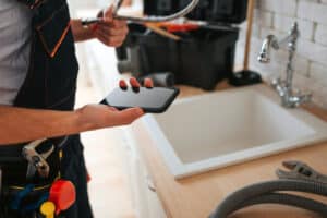 Cut view of man standing in kitchen at sink holding phone and wrench. Hose on desk